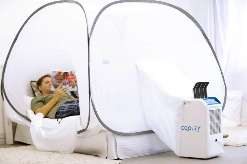 A man lies on a bed reading a magazine inside a white pop-up Igloo Tent with grey-bound hoops, a white Coolzy-Pro standing at the foot of the bed with a fabric sleeve joining it to the tent