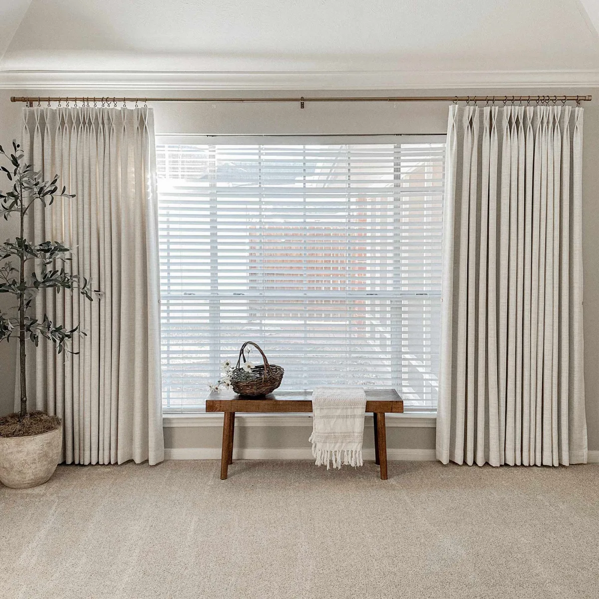 Two off-white pinch-pleat Lesley panels on a brass rod flanking a wide window with white blinds and a wooden bench