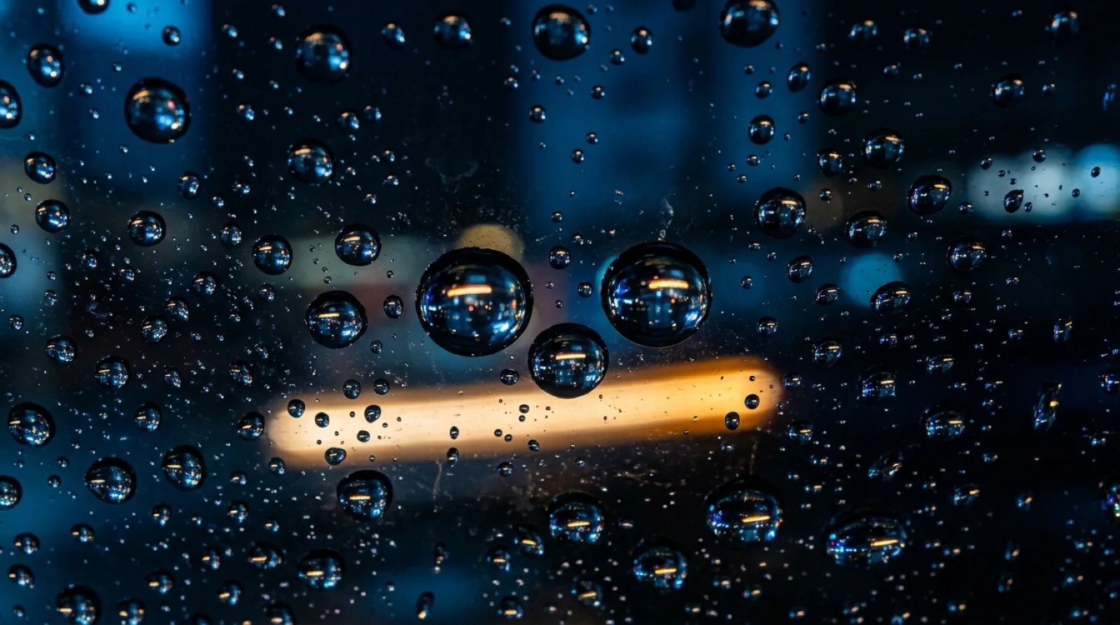 Extreme macro of rain droplets on a windshield at night, each droplet holding a tiny inverted reflection of the glowing street beyond, with one warm amber streak from a passing car