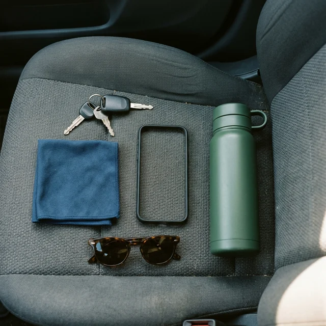 Overhead flat lay on a car seat of keys, a folded microfiber cloth, sunglasses and a steel water bottle arranged around a deliberately empty phone-sized rectangle of space
