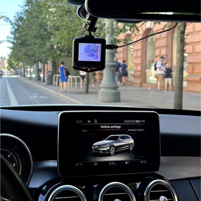 The dash cam mounted behind the rear-view mirror of a car, its screen showing the road ahead while parked on a street