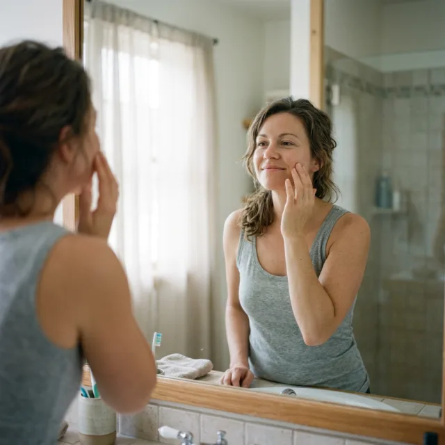 Someone at a bathroom mirror in the morning touching their own cheek with a small satisfied smile, skin looking comfortable rather than tight