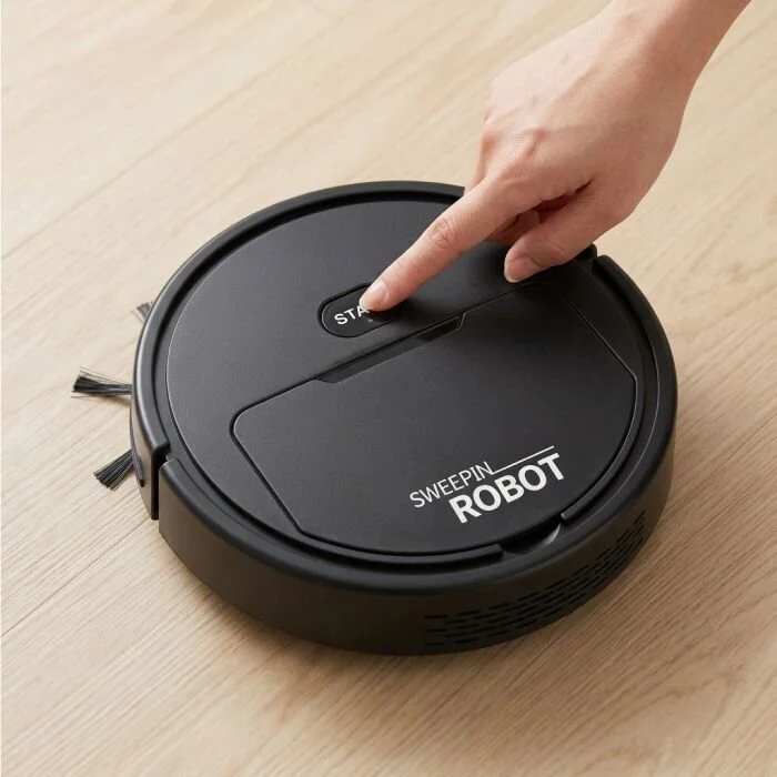 A hand pressing the single start button on top of the DustaPro robot vacuum on a wood floor