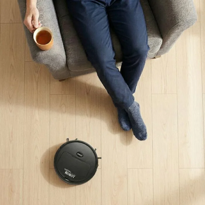 A person relaxing on a sofa with their feet up and a mug of tea while the DustaPro robot vacuum cleans the floor nearby