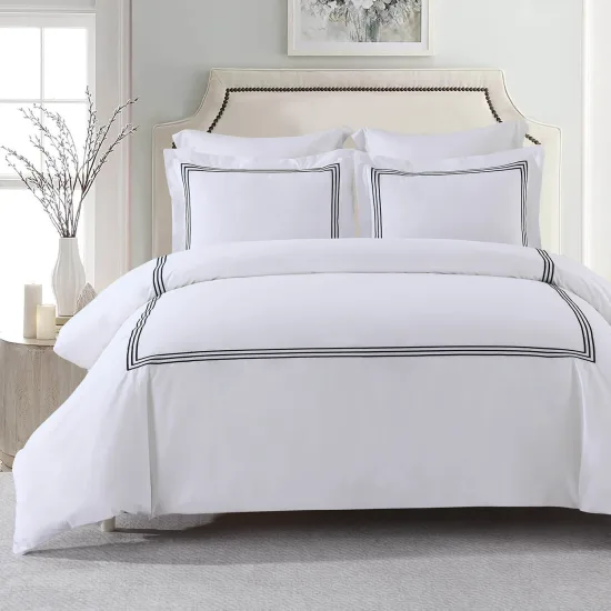 A white duvet cover and two shams with a three-line black embroidered border on a bed with a cream headboard, white flowers on the nightstand