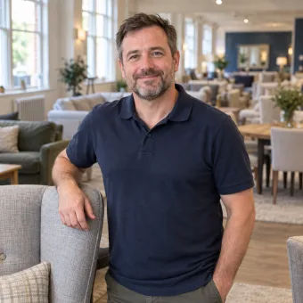 A man in his forties in a plain navy polo shirt standing beside an armchair in a bright furniture showroom