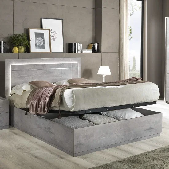 A grey marble effect ottoman bed with a tall headboard lit by an LED strip, the mattress base lifted to show folded bedding stored inside, in a bedroom with a lamp and tall windows