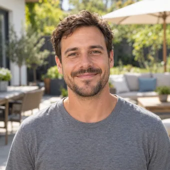 A man in his thirties in a plain grey t-shirt on a sunny patio, garden furniture blurred behind him