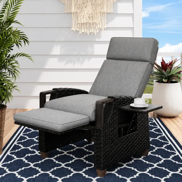 The wicker recliner in Grey with the leg rest raised and a side table holding a cup, on a navy patterned rug beside a white porch wall