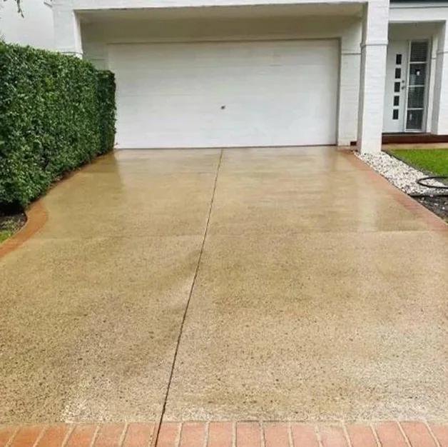 A wet concrete driveway in front of a white house, with a trimmed hedge running down one side