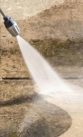 The metal tip at the end of the wand throwing a jet of water at a concrete slab, with water pooling below it