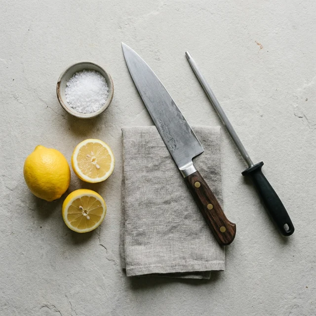 Flat lay on light stone of a folded gray linen towel, a sharpening steel, whole and halved lemons, a small bowl of coarse salt, and a chef's knife laid diagonally