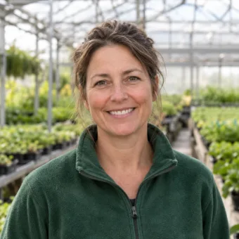 A horticulturist in her forties in a plain green fleece among rows of young plants in a nursery greenhouse