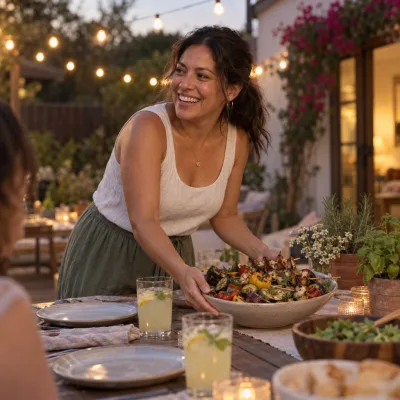 A woman in her thirties laughing as she sets a bowl of grilled vegetables on a patio table under string lights