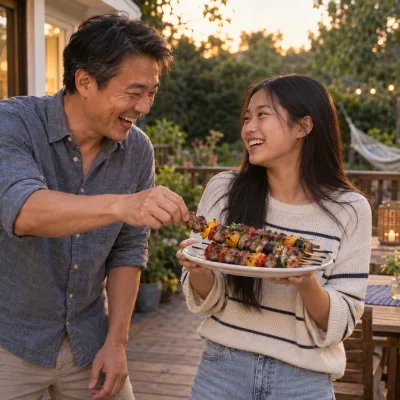 A father and his teenage daughter laughing on a deck as she holds up a plate of grilled kebabs