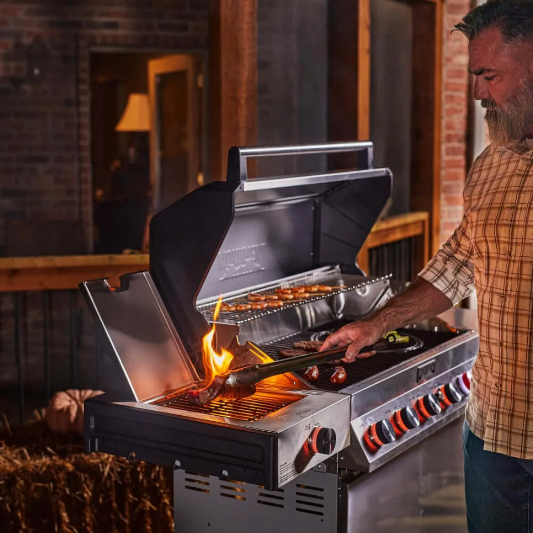 A bearded man in a plaid shirt grilling sausages and vegetables on a Monument Denali 605Pro with the lid open at dusk, a flame rising from the sear burner on the left and red-lit knobs along the front