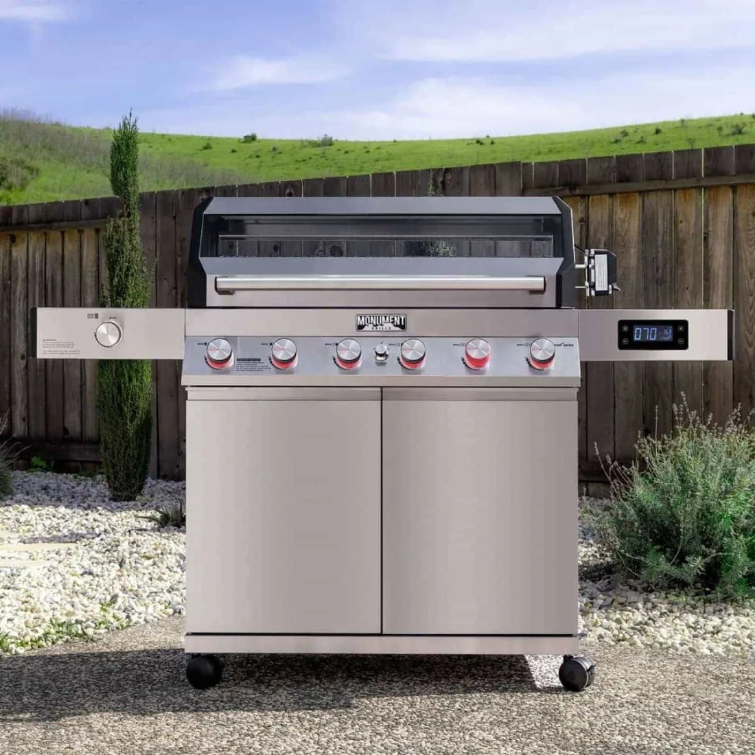A stainless Monument Denali 605Pro gas grill with the lid closed on a stone patio in daylight, a wooden fence and a green field behind it, the digital display lit on the right shelf and a row of knobs across the front