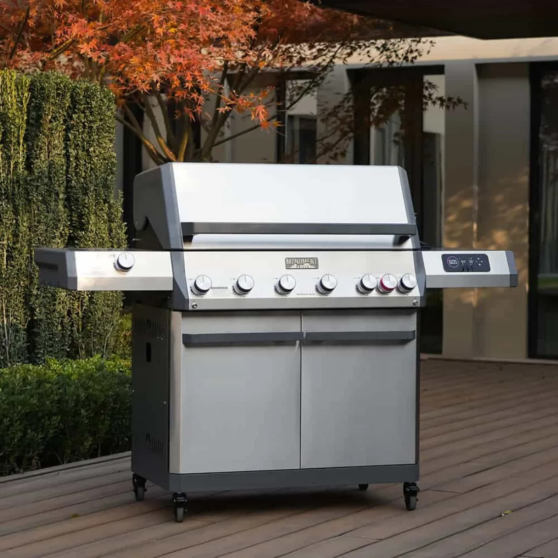 A stainless Monument Eminence 605 gas grill on a wooden deck with a red-leaved maple and a house behind it, lid closed, knobs and a display along the front