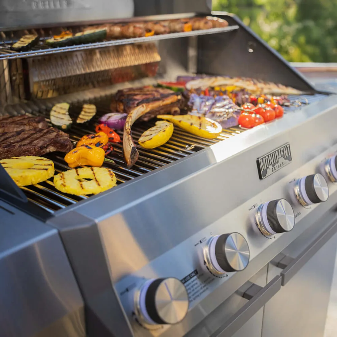 The open Eminence 605 loaded with steaks, corn, peppers, tomatoes and vegetables on stainless grates, a warming rack above and chrome knobs in the foreground