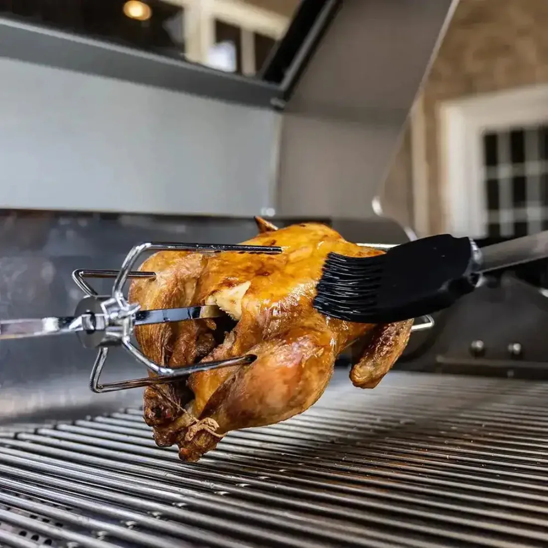 A whole chicken on a rotisserie spit over stainless grates, being brushed with a black silicone basting brush