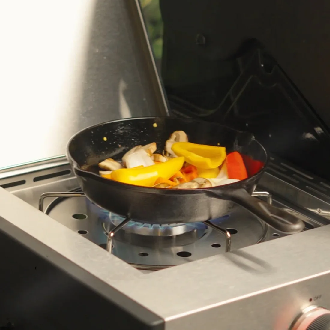 A black skillet of sliced peppers, mushrooms and onions on the side burner of a stainless grill, a blue flame under the pan