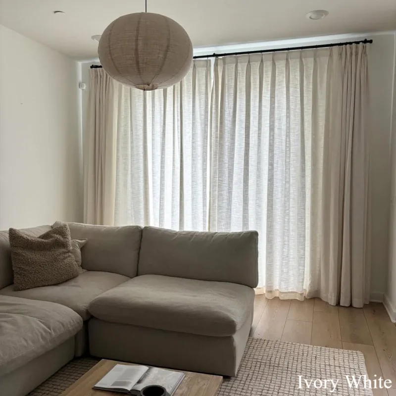 Ivory White pleated linen curtains on a black rod across a wide window behind a beige sectional sofa and a paper lantern lamp