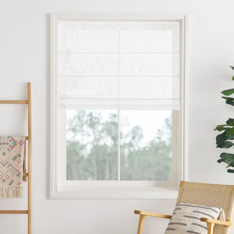 A white Lily Linen roman shade with plain folds lowered halfway in a white-framed window, a wooden ladder with a woven throw and a rattan chair beside it