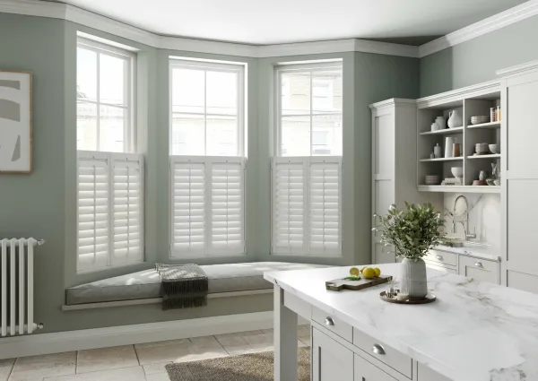 A sage green kitchen bay window with white cafe-style shutters on the lower half of each pane and a cushioned window seat below