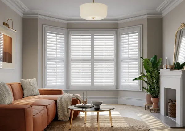 A three-sided bay window with white faux wood shutters, louvres tilted to let the light in, beside a terracotta sofa and a round brass coffee table