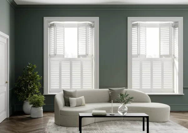 Two windows with white tier-on-tier shutters, the top tier open and the lower tier closed, on a dark green wall behind a curved cream sofa