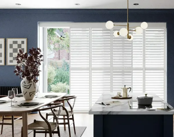 White tracked shutters sliding across a wide garden door in a navy dining room, one panel pushed open to show the trees outside
