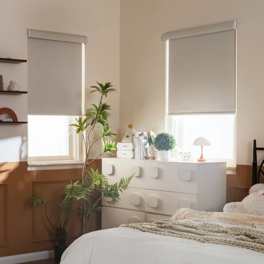 Two light grey SmartWings roller shades lowered about two thirds of the way in a cream and terracotta bedroom, morning light coming in under each shade, a white dresser with a small lamp and a plant between the two windows