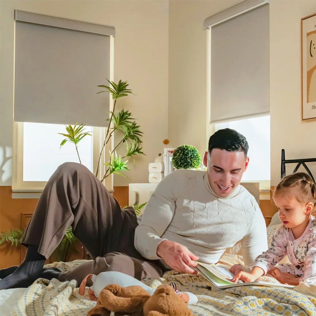 A man in a cream sweater and a small girl in floral pajamas reading a picture book on a bed, two grey roller shades lowered behind them, a plush bear in the foreground