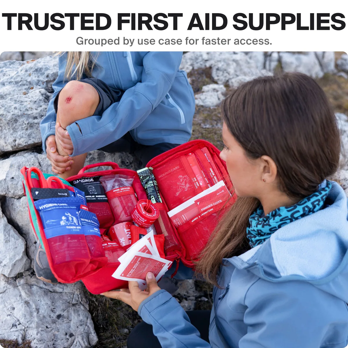 A woman on a rocky trail opening the red kit beside a child with a scraped knee, the seller's own photo