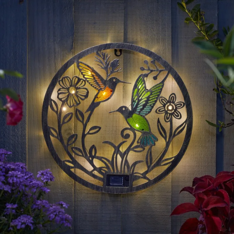 A round metal Solar Hummingbird Wings Decor Disc on a grey fence, lit warm white, with two stained-glass hummingbirds and cut-out flowers