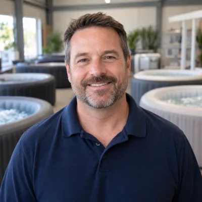 A man in his forties in a plain navy polo shirt, round inflatable hot tubs blurred in a bright showroom behind him