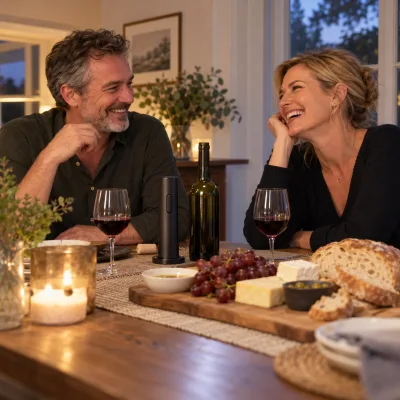 A couple in their fifties laugh at their dining table over a plate of cheese and bread, a slim black electric wine opener standing beside an opened bottle and two glasses